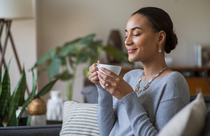 Frau sitzt auf einem Sofa, hat die Augen geschlossen und einen entspannten Gesichtsausdruck. Sie lächelt. Sie hält in ihren Händen eine Tasse Kaffee oder Tee.